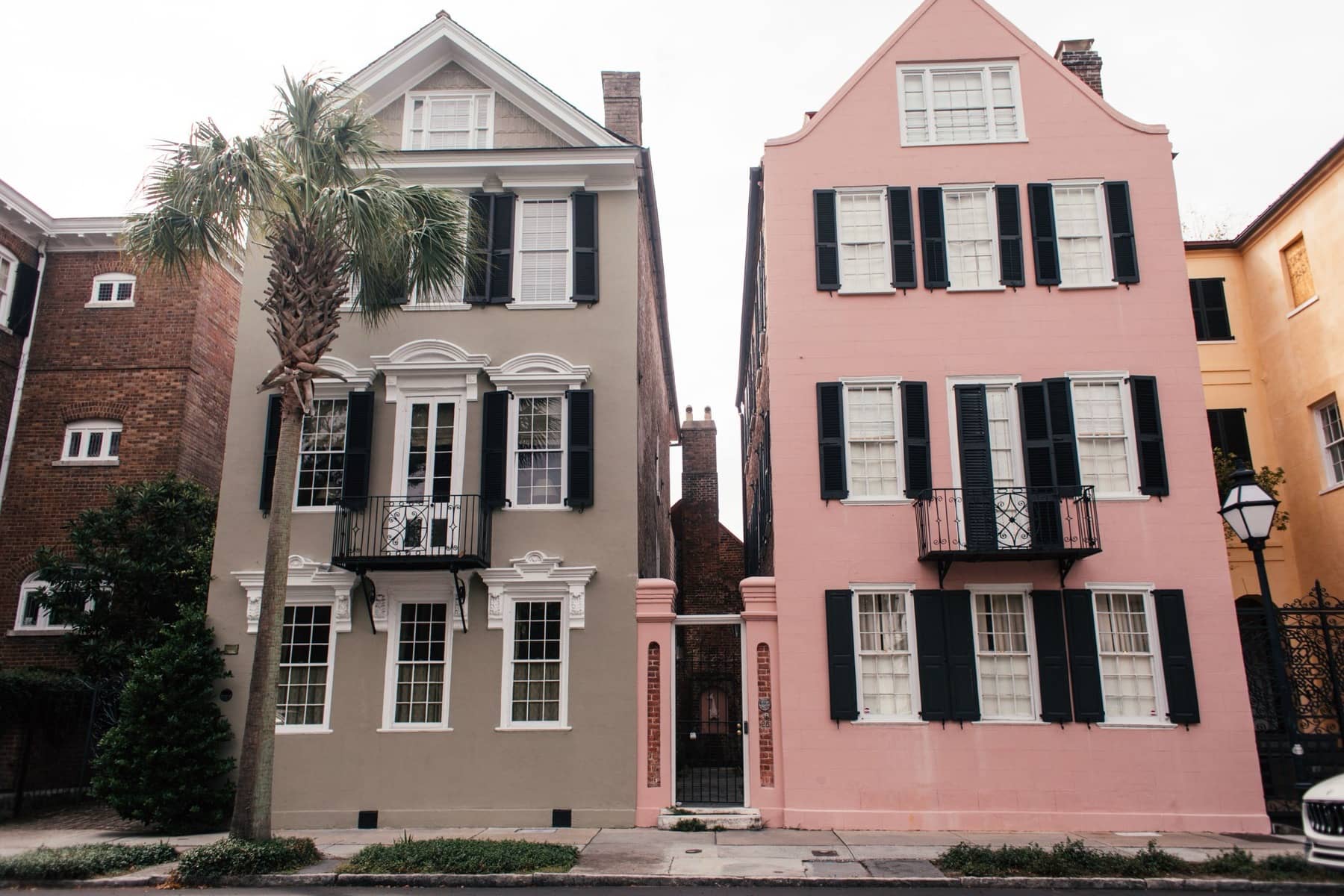 Two historic three-story houses, one gray and one pink, with black shutters, iron balconies, and a palmetto tree on a downtown street, Charleston area